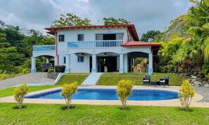 Una casa de dos pisos con piscina, un patio cubierto y vistas de la vegetación circundante.
