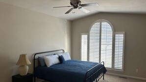 Un dormitorio con cama, ventilador de techo, lámpara y ventana con persianas.