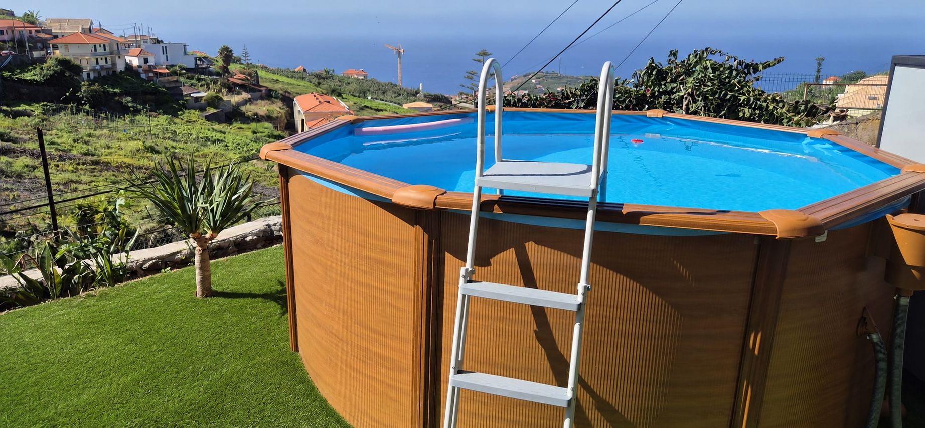 Piscina elevada de madera con escalera, rodeada de césped y vistas a casas y colinas.