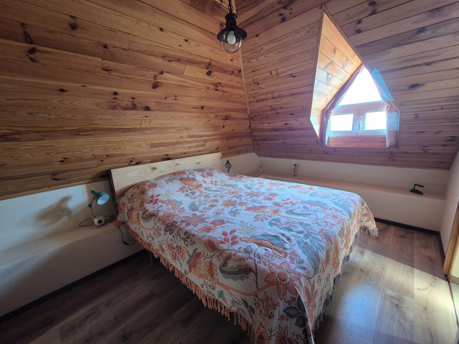 Un dormitorio con techo de madera, una cama con colcha estampada, una mesita de noche, una lámpara y un ventanal con techo triangular.