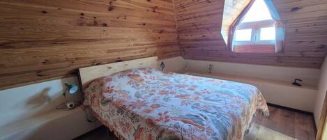 Un dormitorio con techo de madera, una cama con colcha estampada, una mesita de noche, una lámpara y un ventanal con techo triangular.