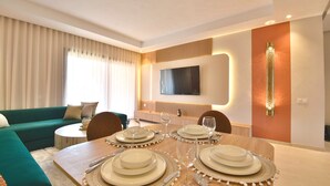 A modern living room with a dining area, a sofa, and a TV mounted on the wall. - Modern 2BR/2BA Apt • Pool View Terrace • Fast Fiber WiFi • Noria Resort (Marrakech)