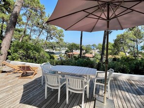 A wooden deck with a table and chairs, an umbrella, and a view of a coastal town. - Vacation home with wonderful terrace and far-reaching views to the bay (Lège-Cap-Ferret)