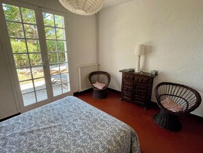 Une pièce avec une grande fenêtre, un lit, une commode et deux fauteuils en osier.