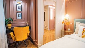 A hotel room with a bed, a desk with a chair, a small table with a lamp, and a door leading to another room. - Pink Perfection -  1 Bedroom Designer Apartment in Bucharest (București)