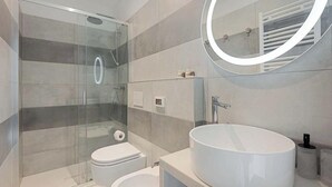 Uma casa de banho moderna com chuveiro envolto em vidro, sanita branca e uma grande lavatório redondo.