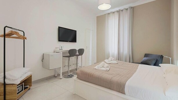 Quarto de hotel com cama, escrivaninha com duas cadeiras, televisão, guarda-roupa e janela com cortinas.