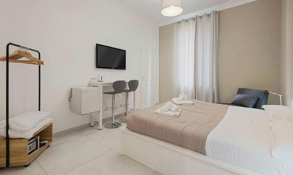 Quarto de hotel com cama, escrivaninha com duas cadeiras, televisão, guarda-roupa e janela com cortinas.