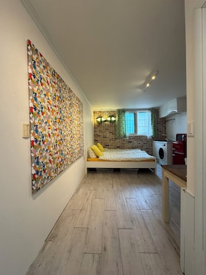 Ein kleines Schlafzimmer mit Schlafbox, farbenfroher Wandkunst, Backsteinwand, Fenster mit Vorhängen und einer Waschmaschine.