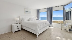A bedroom with a large bed, a nightstand, a chair, and a view of the beach. - Surf & Racquet B170 - Hosted by Moore AI Rentals (Fernandina Beach)