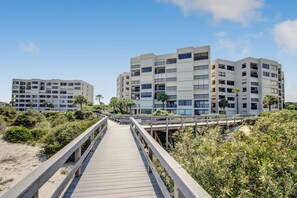 A wooden boardwalk leading to a modern multi-story building with balconies, surrounded by greenery and sand. - Surf & Racquet B170 - Hosted by Moore AI Rentals (Fernandina Beach)