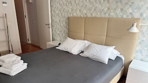 Een keurig opgemaakt bed met witte kussens en een grijze plaid, een nachtkastje met een lamp, en een muur met een geometrisch patroon.