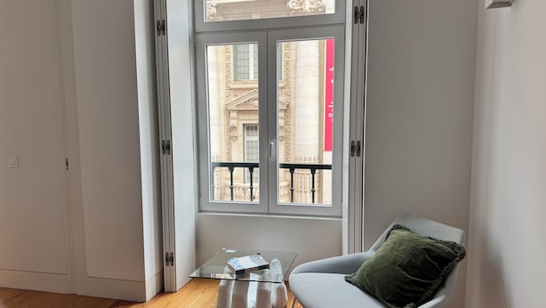 Een hoekappartement met een glazen tafel, een grijze stoel met een groen kussen, en uitzicht op een gebouw door een raam.