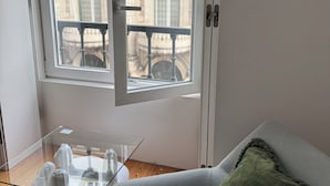 Een kamer met een tafel met glazen blad, een grijze bank en uitzicht op een gebouw met kolommen en een balkon.