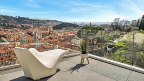 A modern balcony with a white chair and a potted plant, overlooking a cityscape with red-roofed buildings and a distant view of the sea.