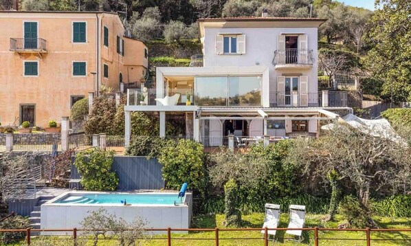 Uma casa moderna de dois andares com piscina, cercada por vegetação e outros edifícios.