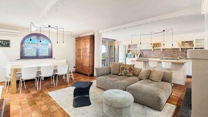 A modern kitchen with white cabinets, a dining area with a white table and chairs, and a living room with a sofa and ottoman.
