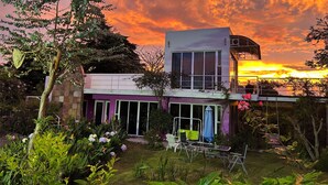 Una casa moderna de dos pisos con balcón, rodeada de abundante vegetación y plantas en flor.