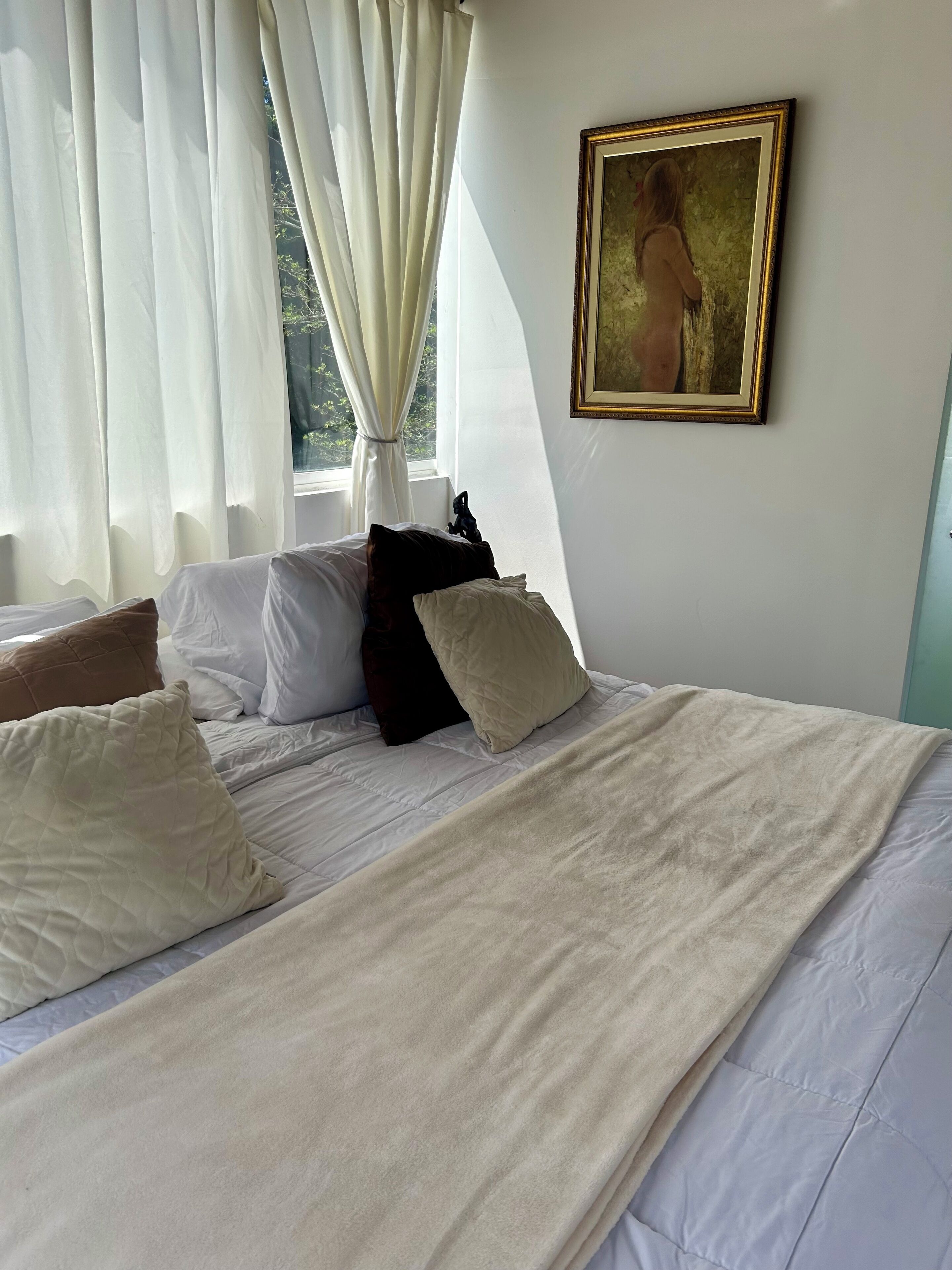Un lit bien fait, avec une literie blanche et beige, un tableau accroché au mur et des rideaux légers à la fenêtre.