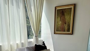 Una cama bien tendida con ropa de cama blanca y beige, un cuadro en la pared y cortinas translúcidas en la ventana.
