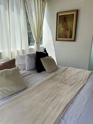 Una cama bien tendida con ropa de cama blanca y beige, un cuadro en la pared y cortinas translúcidas en la ventana.