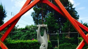 Eine Skulptur einer Hand, die ein Herz hält, umgeben von einem roten, geometrischen Rahmen, vor einer grünen Wiese mit Bäumen und einem Zaun im Hintergrund.