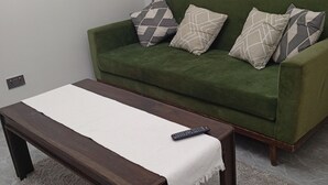 Un sofá verde con cojines estampados, una mesa de centro de madera con un mantel blanco y un control remoto encima.