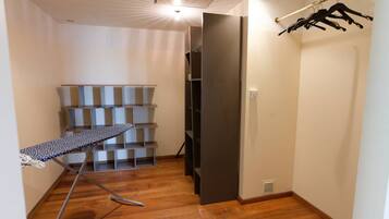 Una habitación con piso de madera, una tabla de planchar de metal, estantes y un perchero.