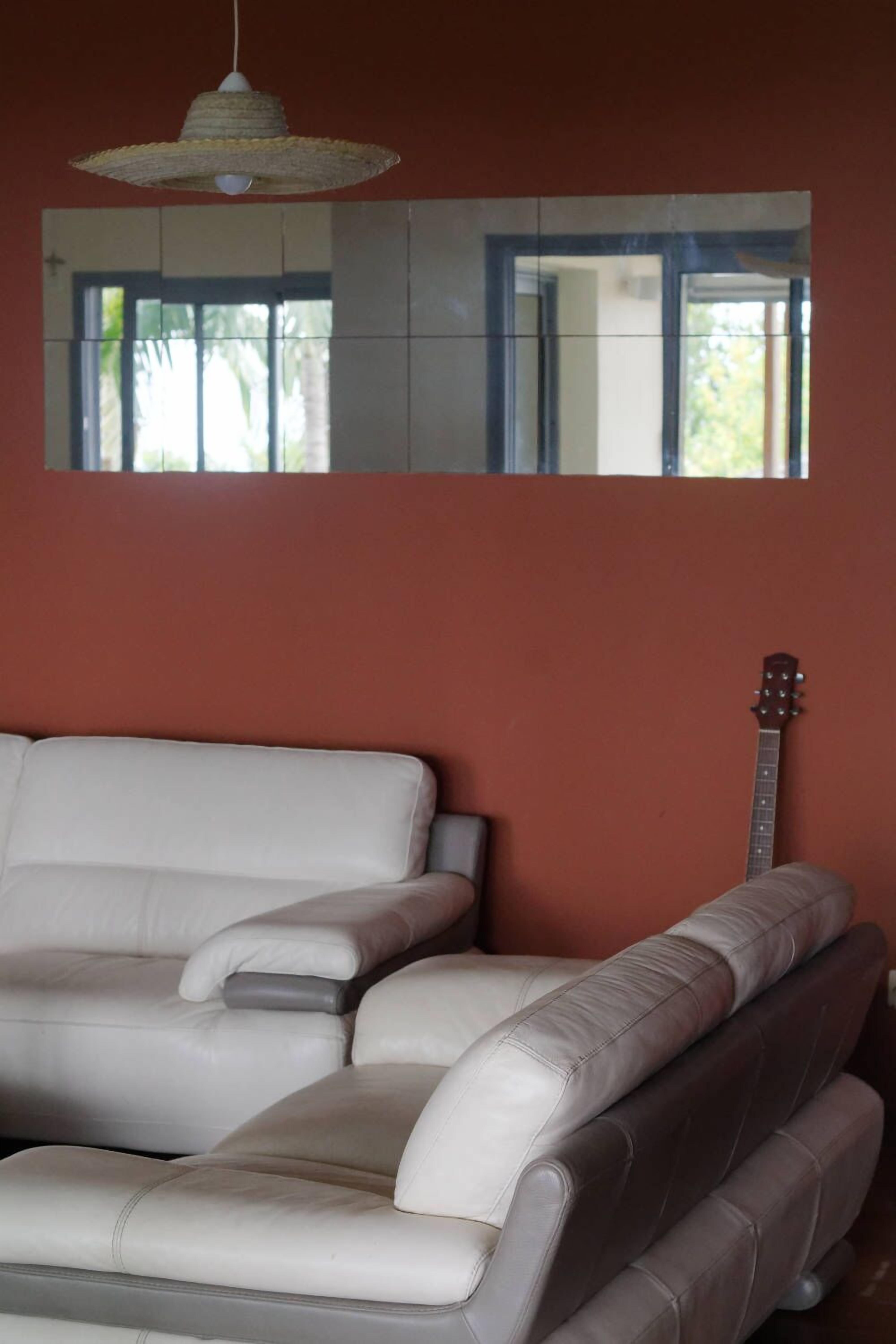 A room with a white leather sofa, a guitar leaning against the wall, and a large mirror reflecting a window with greenery outside.