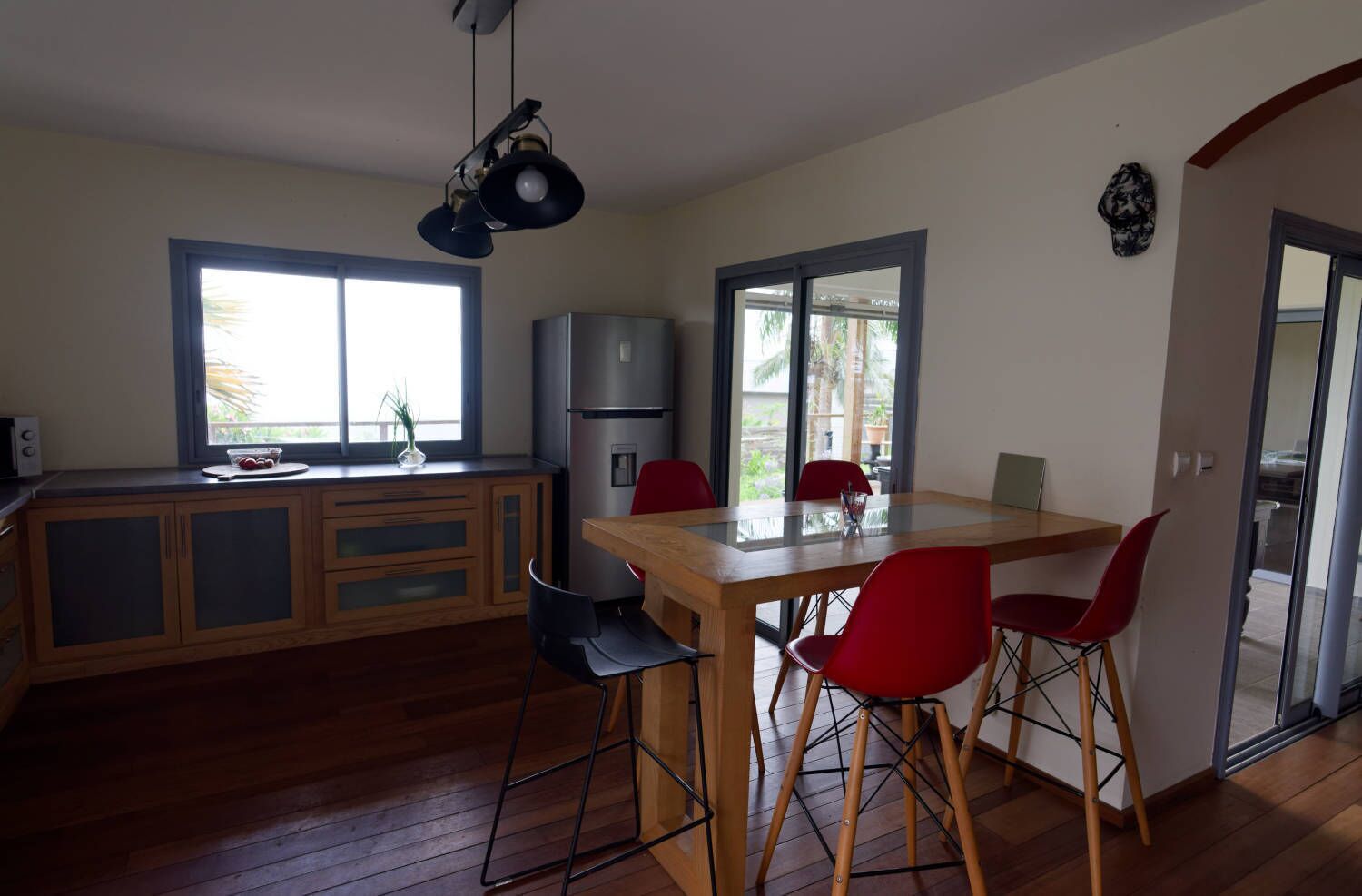A modern kitchen with wooden cabinets, a stainless steel refrigerator, and a dining area with a glass table and red chairs.