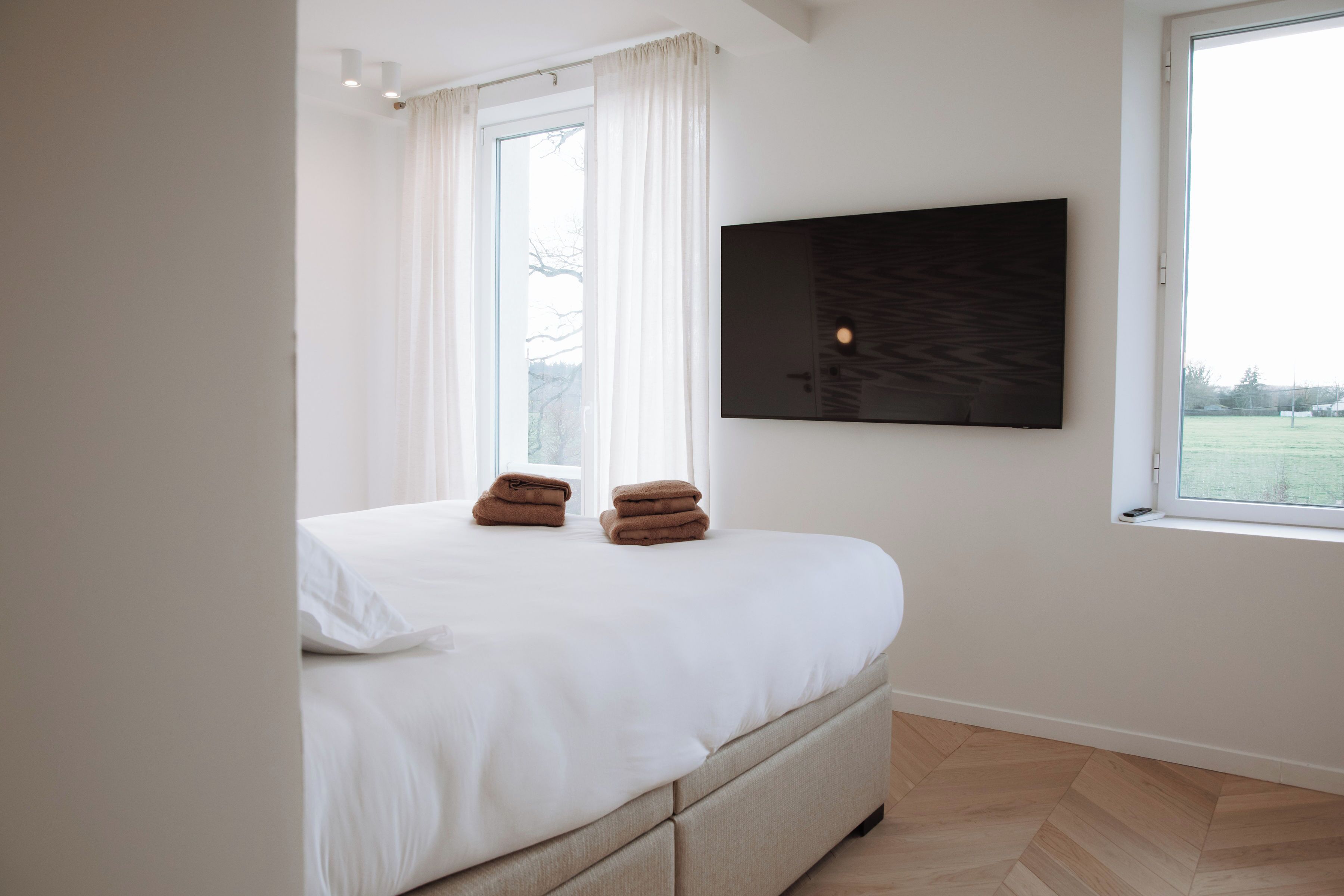 A bedroom with a bed, a flat-screen TV mounted on the wall, and a window with curtains.