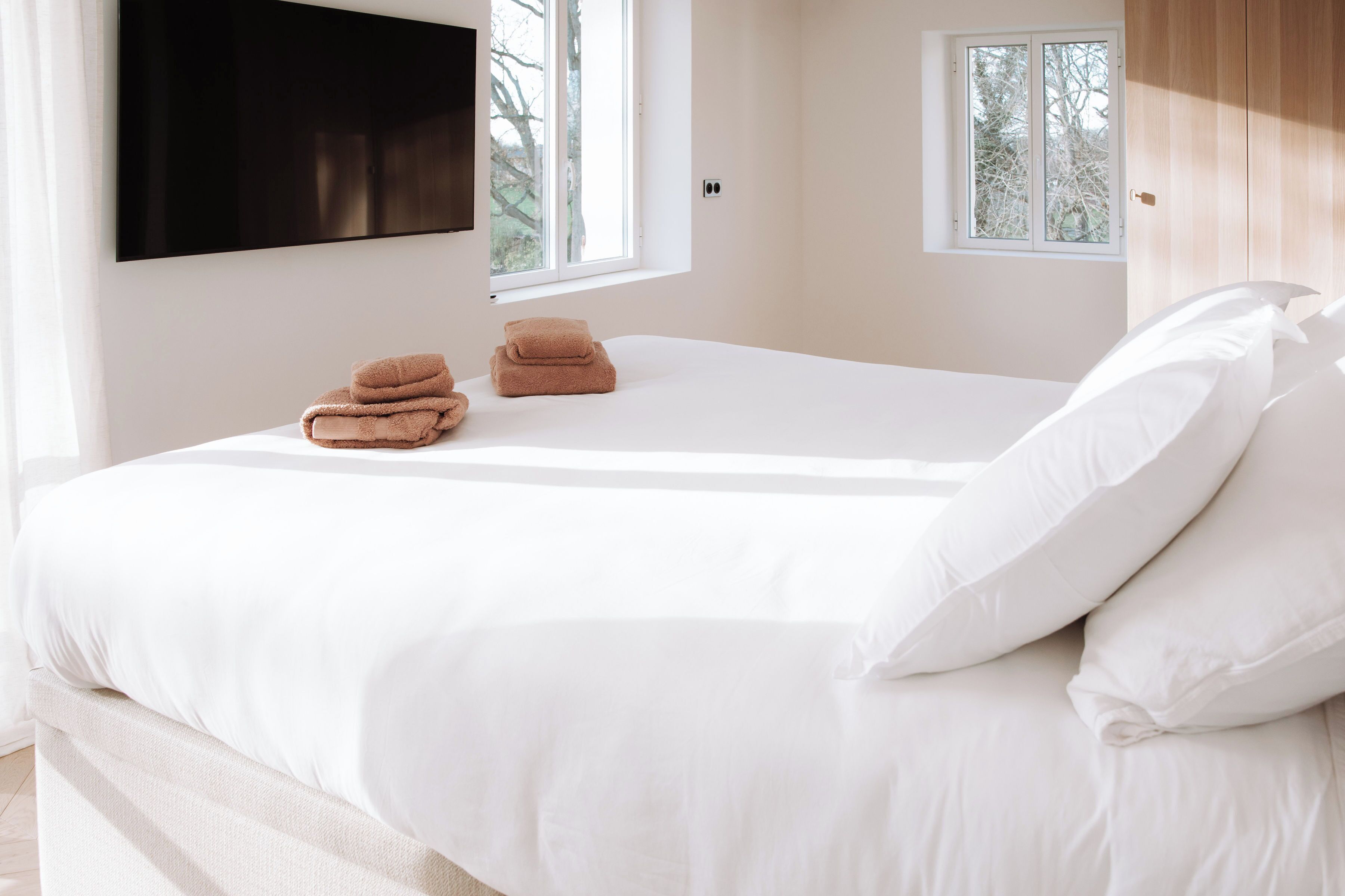 Un dormitorio con una cama, un televisor montado en la pared y una ventana con vistas a árboles.