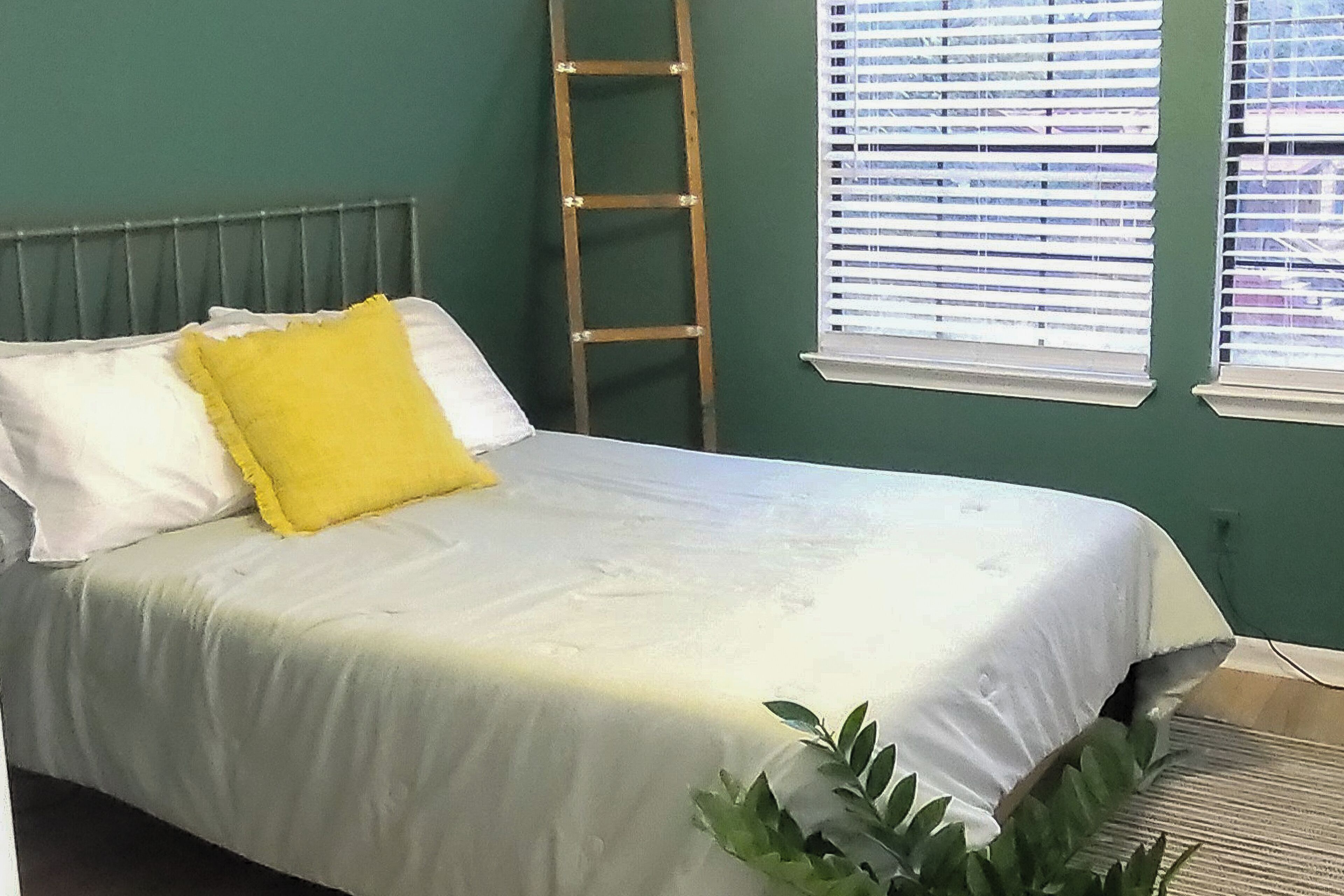 Una cama con almohadas blancas y amarillas, una escalera de madera y una ventana con persianas.