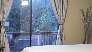 Une chambre avec une grande fenêtre, des rideaux, un lit et une vue sur une terrasse et la verdure environnante.