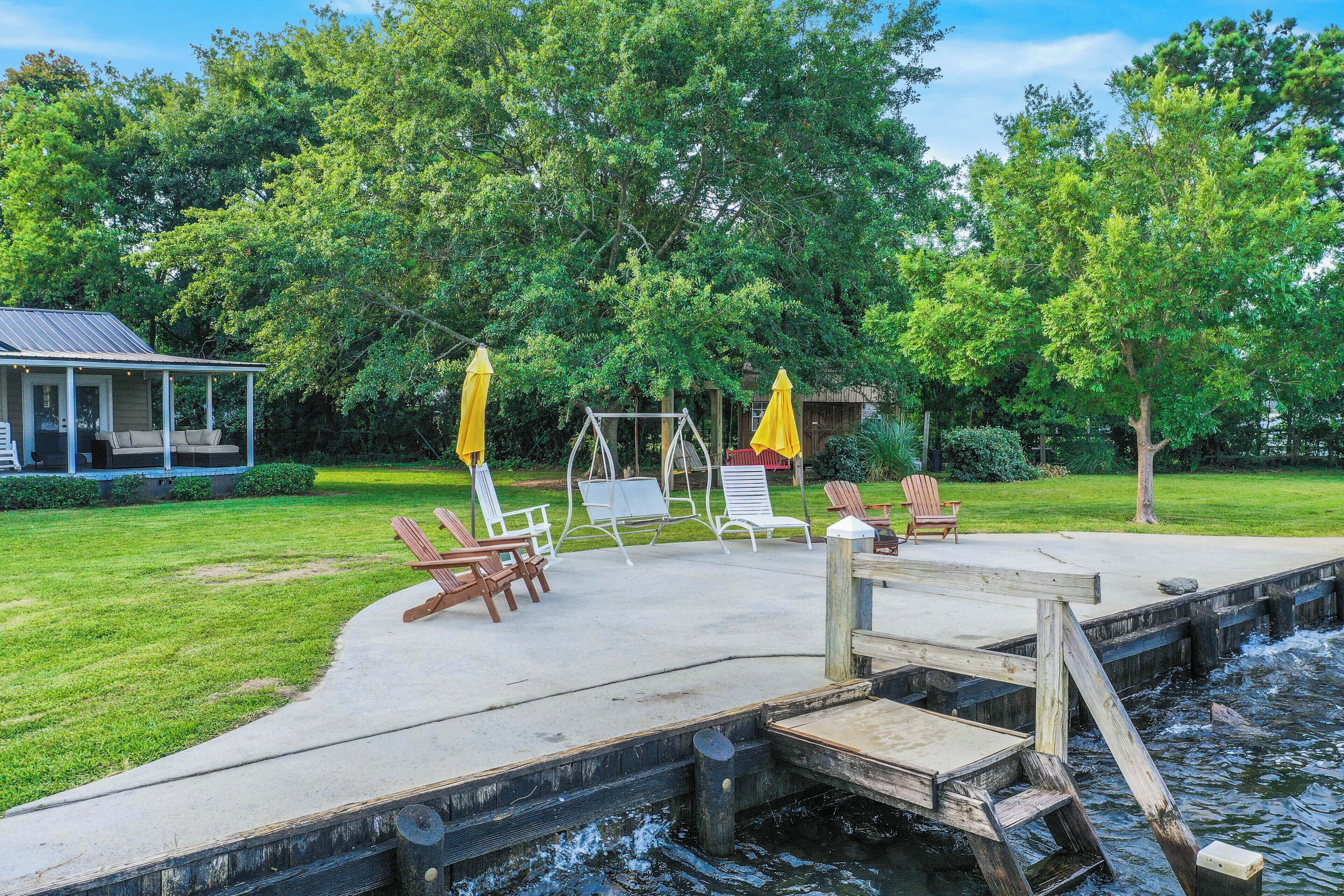 A concrete deck with a wooden dock extending into a body of water, surrounded by lush greenery and outdoor seating.