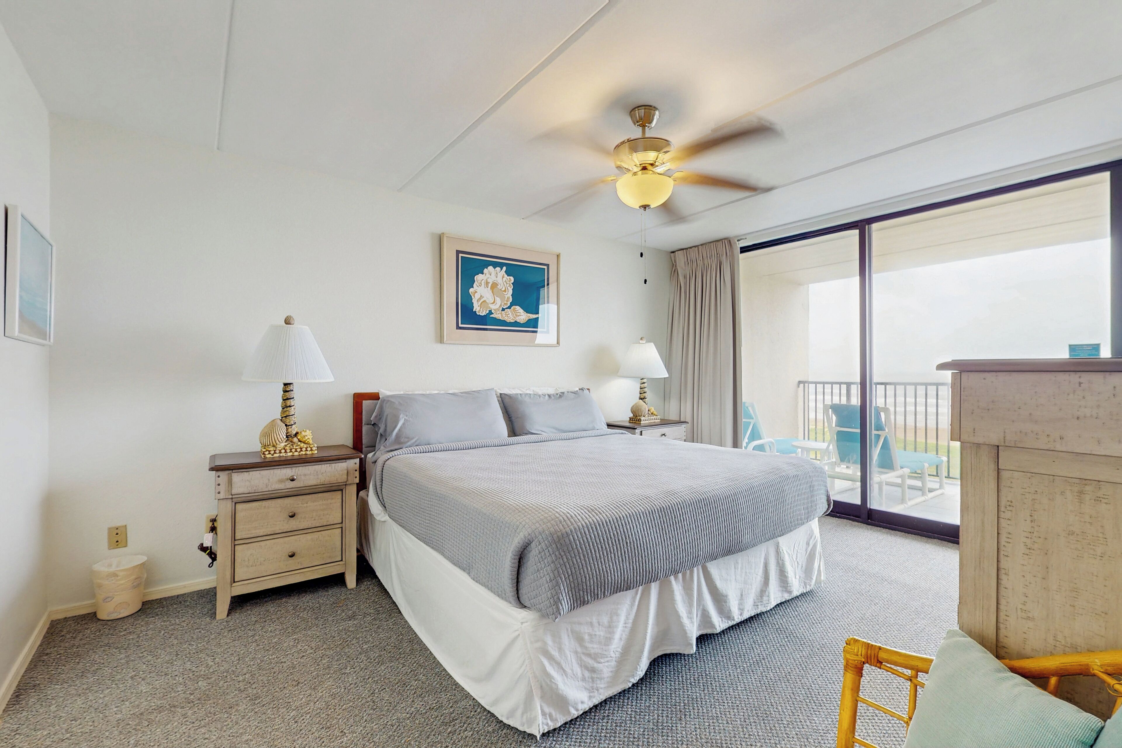 A hotel room with a bed, two bedside tables with lamps, a ceiling fan, a large window with a view, and a framed picture on the wall.