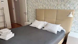 A neatly made bed with white pillows and a grey bedspread, a bedside table with a lamp, and a wall with a geometric pattern. - The Crucifixo Collection - Serene (Lisboa)