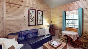 A cozy cabin interior with a wooden wall, a leather sofa, a small table, and a window with curtains. - Serene & Secluded Paige Cabin: Dogs Welcome! (Paige)