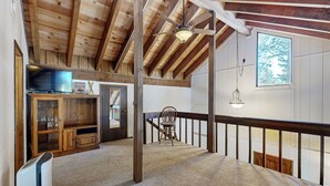 Una habitación amplia con techo alto, vigas de madera, ventilador de techo, un gran ventanal con vistas, un comedor con una silla y un televisor sobre un mueble.