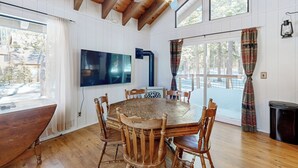 Un comedor con una mesa y sillas de madera, un televisor de pantalla plana y ventanas amplias con cortinas.