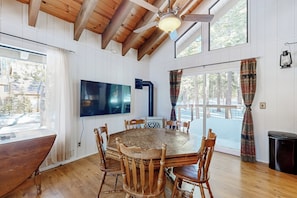 Ein Holzesstisch mit sechs Stühlen, einem Flachbildfernseher und einem Deckenventilator in einem Raum mit weißen Wänden und Holzbalken.
