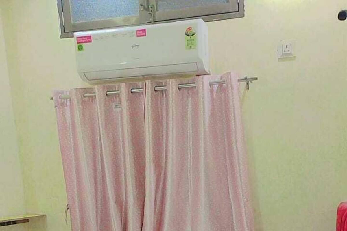 A small, single-bed room with a wooden headboard, a window with a metal grill, and a wall-mounted air conditioner.
