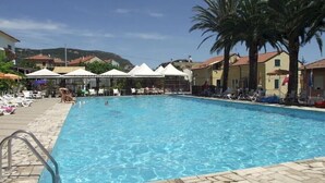 A swimming pool with lounge chairs and umbrellas, surrounded by buildings and palm trees. - Al Borgo Village (Pietra Ligure)