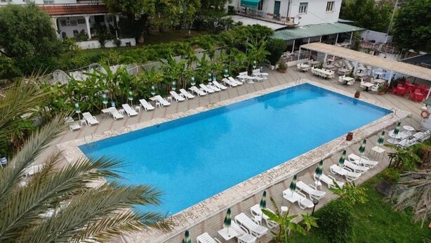A rectangular swimming pool with white lounge chairs and umbrellas arranged around it, surrounded by greenery and a building in the background. - Al Borgo Village (Pietra Ligure)