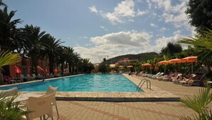 A swimming pool with lounge chairs and umbrellas, surrounded by palm trees and a clear sky. - Al Borgo Village (Pietra Ligure)