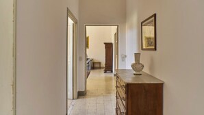 Un couloir étroit, dans lequel on trouve une commode en bois et un vase posé dessus, mène à une pièce où se trouvent un lit et une chaise.