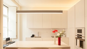 A modern kitchen with white cabinetry, a central island with two chairs, a red vase with flowers, and a built-in oven. - FLH Downtown Central Minimalist Apartment (Lisboa ciudad)
