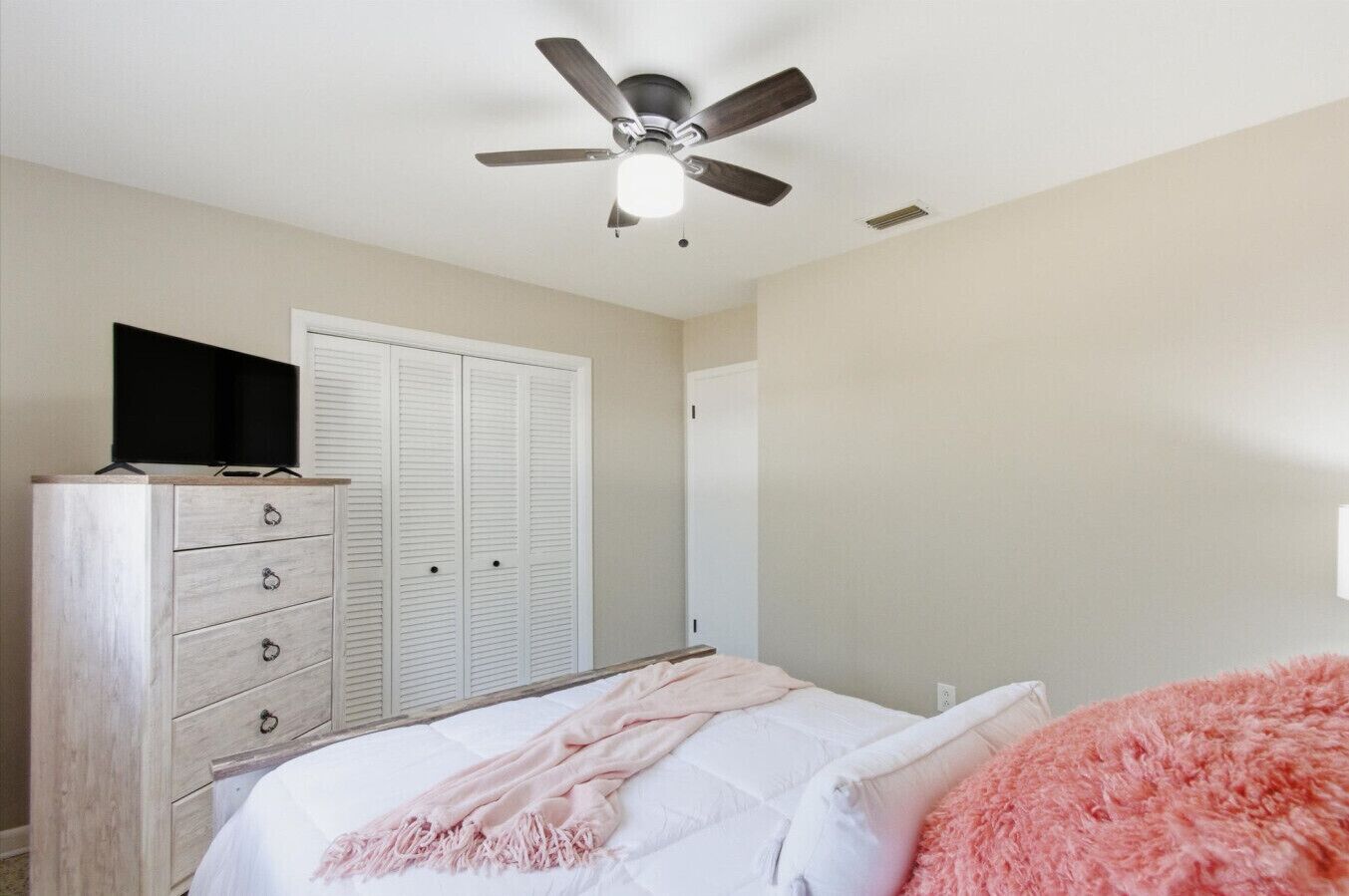 Quarto com cama, penteadeira, televisão, guarda-roupa e ventoinha no teto.