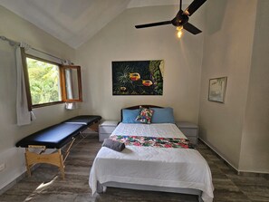 Ein Schlafzimmer mit Bett, Sitzbank, Deckenventilator, Fenster mit Vorhängen und Wandbild.