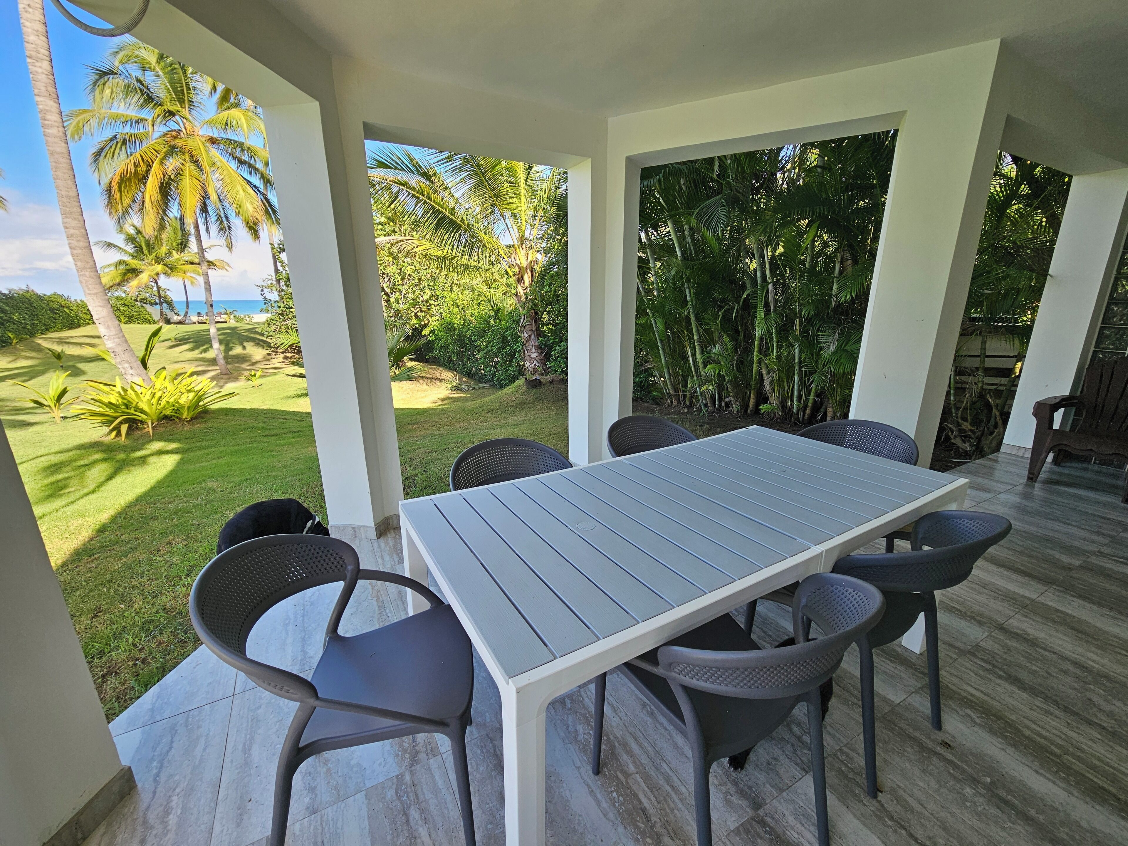 A covered outdoor dining area with a table and chairs, surrounded by lush greenery and palm trees.
